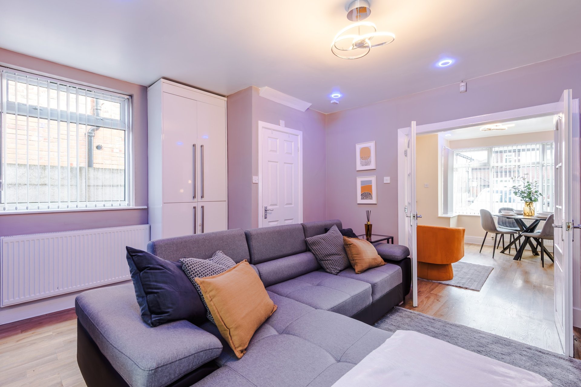 Modern living room with purple walls, gray corner sofa, orange and blue cushions, wooden floor