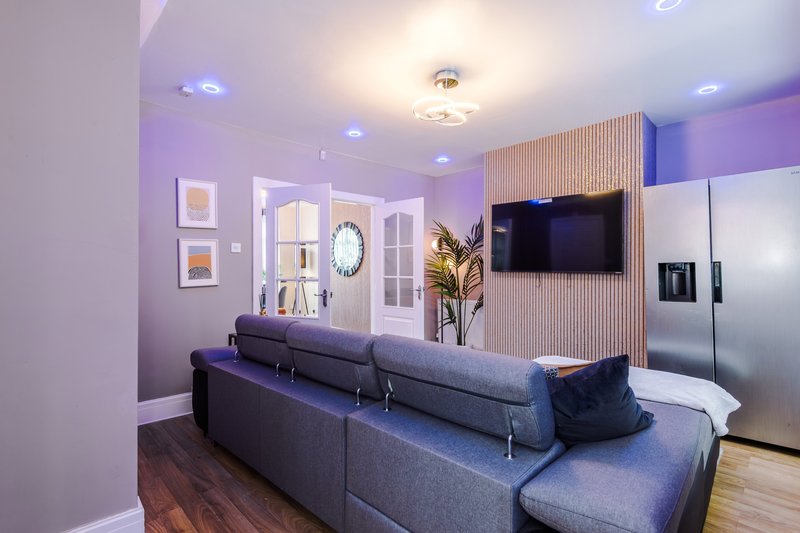 Modern living room with grey corner sofa, wall-mounted TV, purple accent walls and stainless steel kitchen appliances