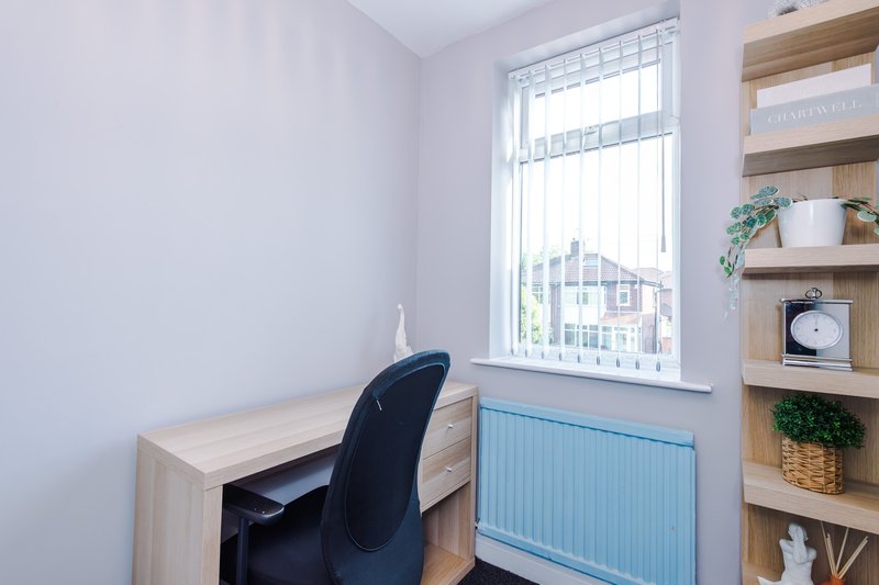 Small bedroom office with desk, black chair, wooden shelving and window overlooking street