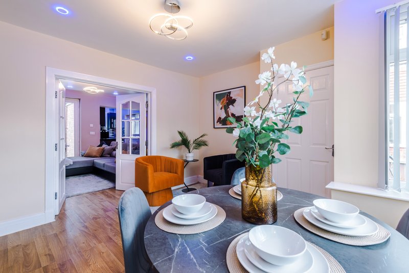 Open-plan dining and living area with table set for three, orange chair, flowers, and purple-lit room beyond