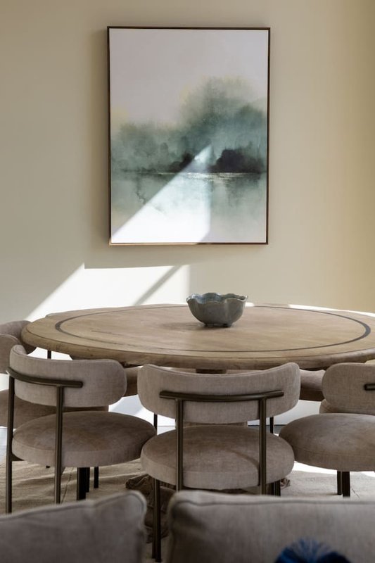 Modern dining area with round wooden table, upholstered chairs, and abstract landscape artwork on wall