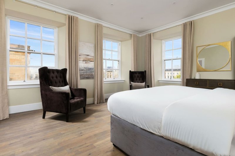 Spacious bedroom with large bed, dark wingback chair, multiple windows with city views