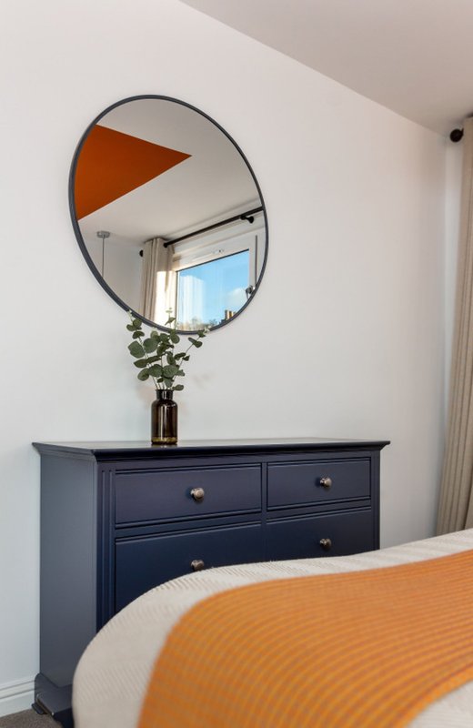 Bedroom with navy dresser, round mirror, potted plant, and orange bedding detail