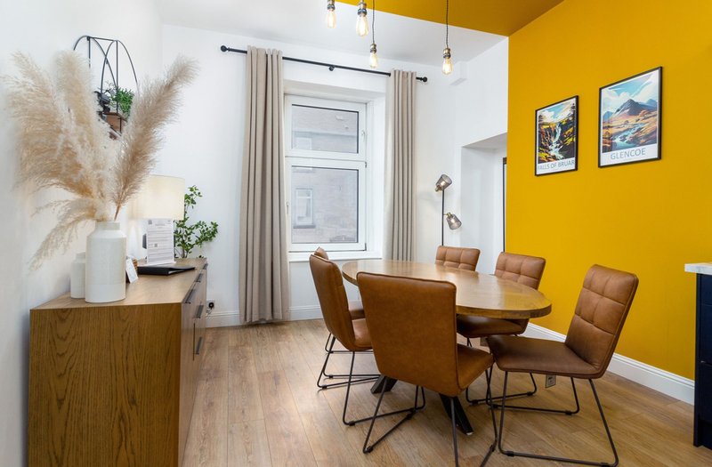 Bright dining area with wooden table, tan leather chairs, yellow accent wall, and framed artwork