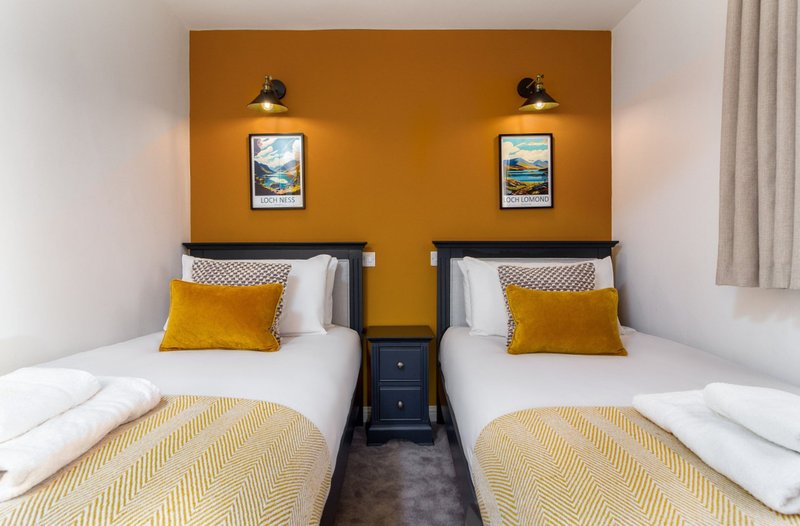 Twin bedroom with two beds, yellow mustard headboard wall, framed landscape artwork, and matching bedside lamps