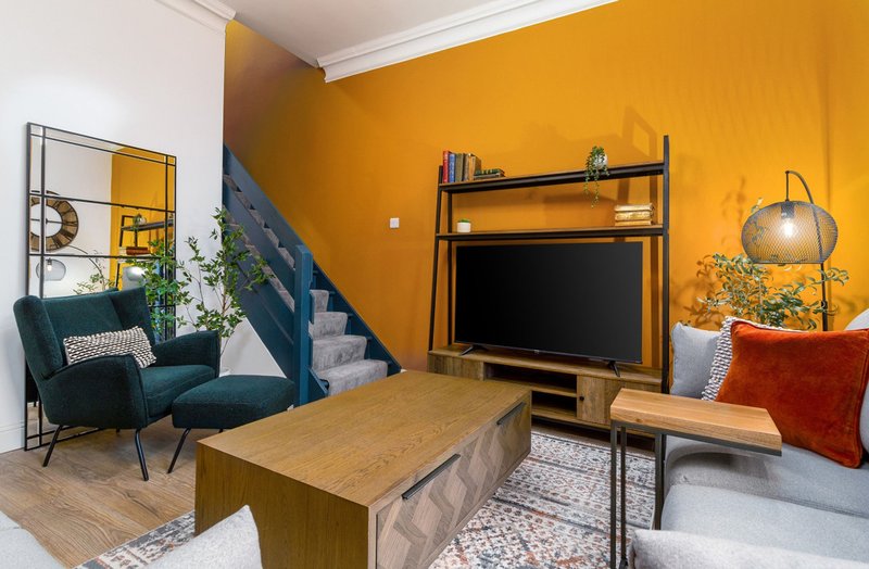 Living room with wooden coffee table, teal armchair, TV unit, orange accent wall and stairs