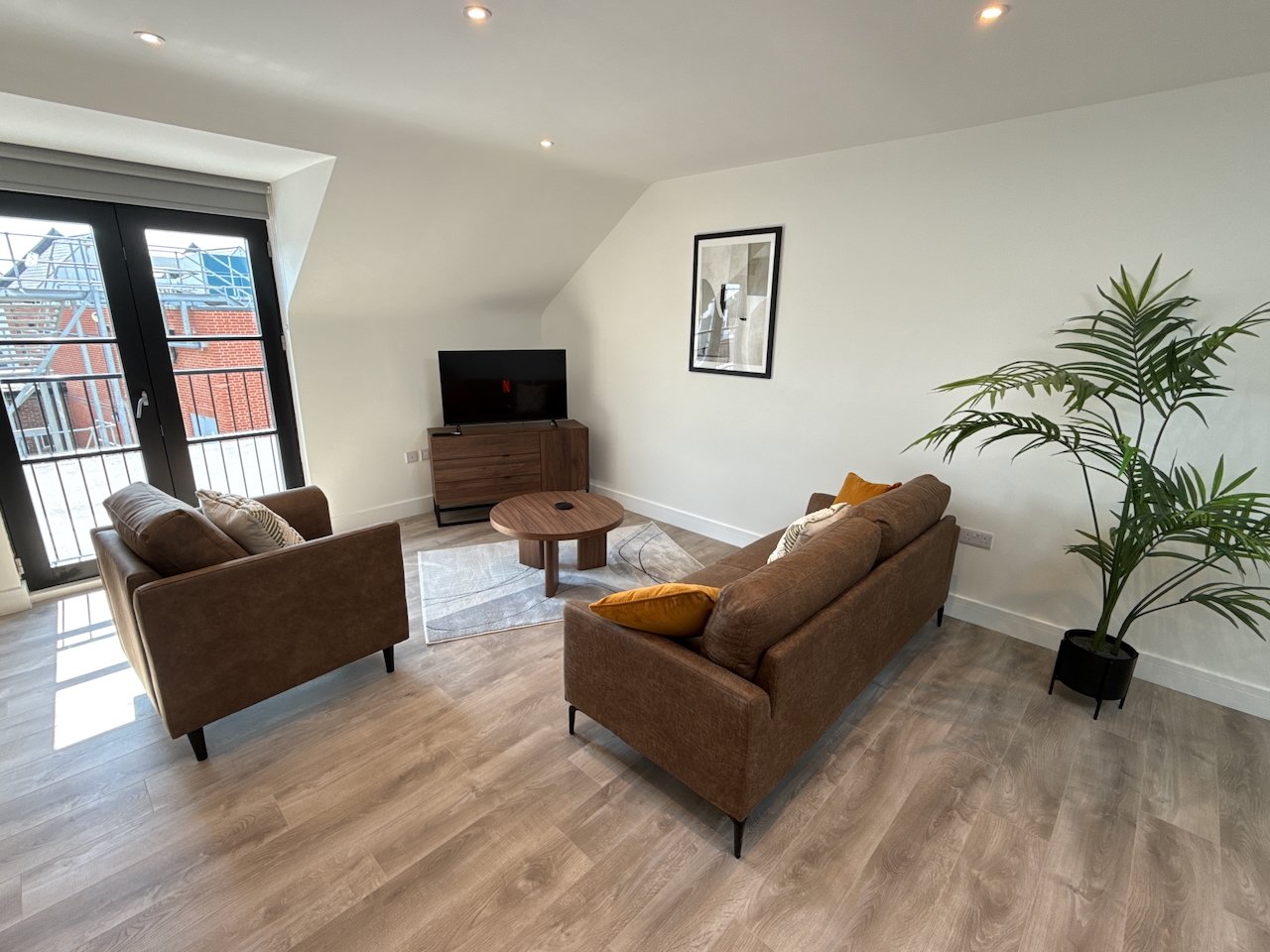 Modern living room with two brown sofas, wooden floor, TV, plant and balcony access