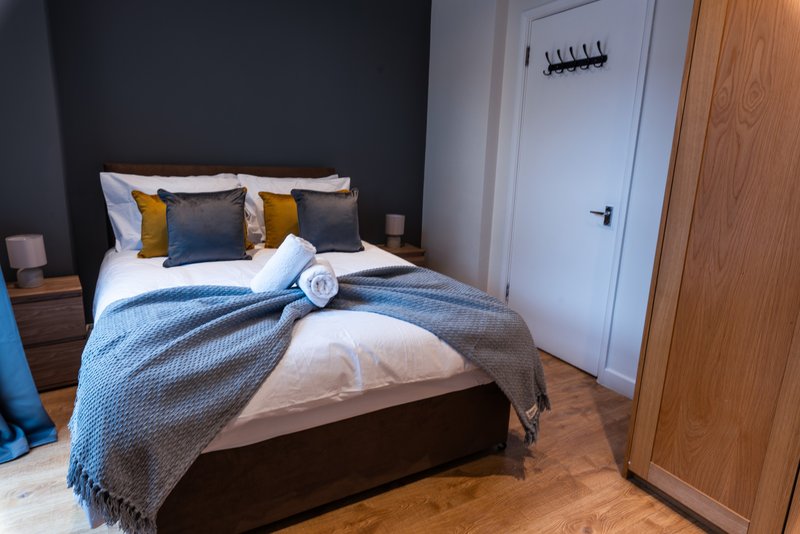 Modern bedroom with double bed, blue bedding, gold and navy cushions, wooden flooring and dark feature wall