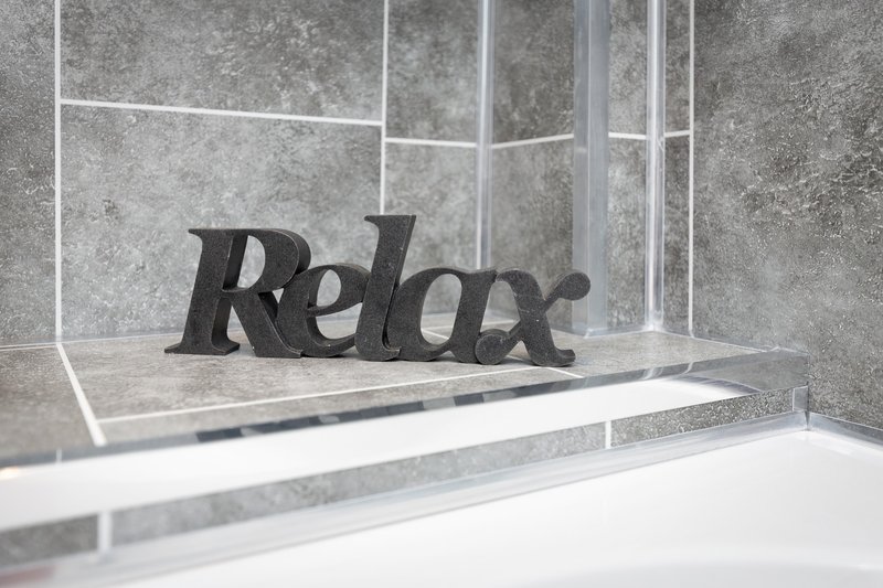 Modern bathroom with gray tile walls, white bathtub edge, and black decorative 'Relax' word ornament