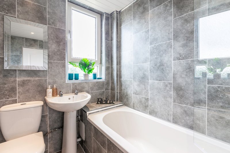Modern bathroom with grey tile walls, pedestal sink, toilet, bathtub and window