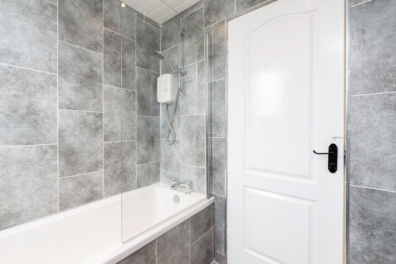 Modern bathroom with grey tile walls, white bathtub, glass shower screen and white door