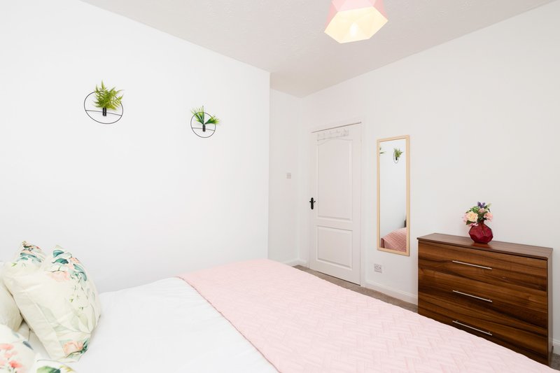 Bright bedroom with double bed, pink bedding, wooden dresser, wall plants, and mirror