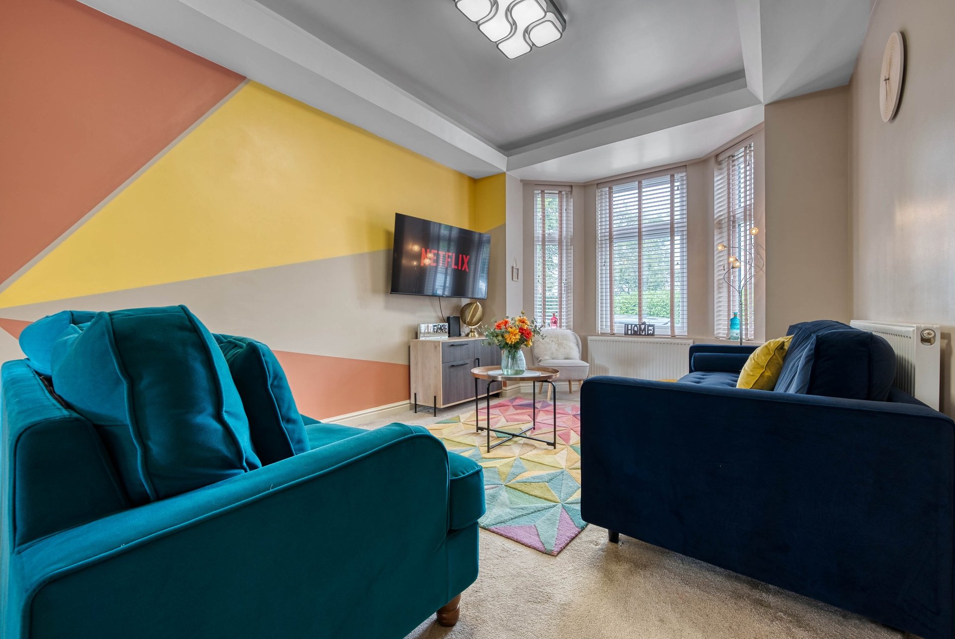 Colorful living room with teal and navy sofas, geometric yellow wall, mounted TV, and large windows