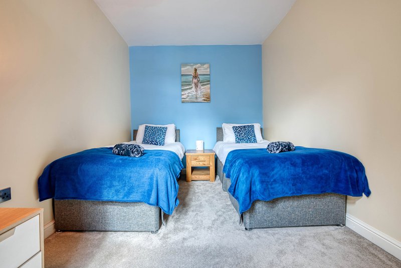 Twin bedroom with two blue beds, wooden nightstand, light carpet and blue accent wall