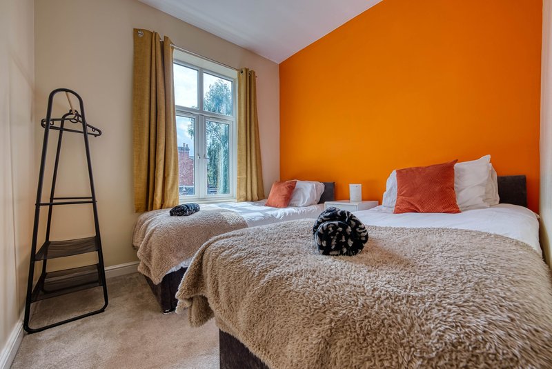 Bright bedroom with two beds, orange accent wall, window with curtains, and decorative cushions
