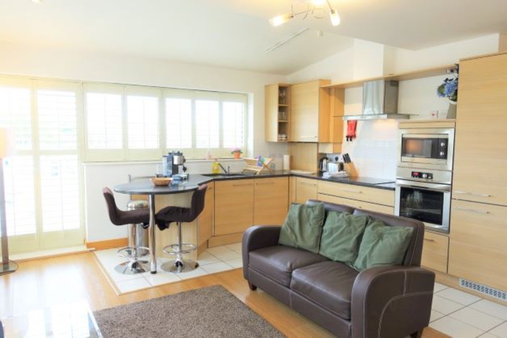 Open-plan kitchen and living area with wooden cabinetry, brown leather sofa, and large windows