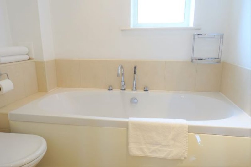 Modern bathroom with large white bathtub, cream tile surround, toilet, and window