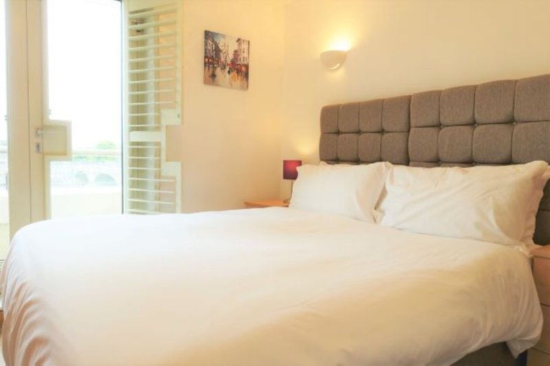 Bedroom with double bed, grey upholstered headboard, white bedding, and window with blinds