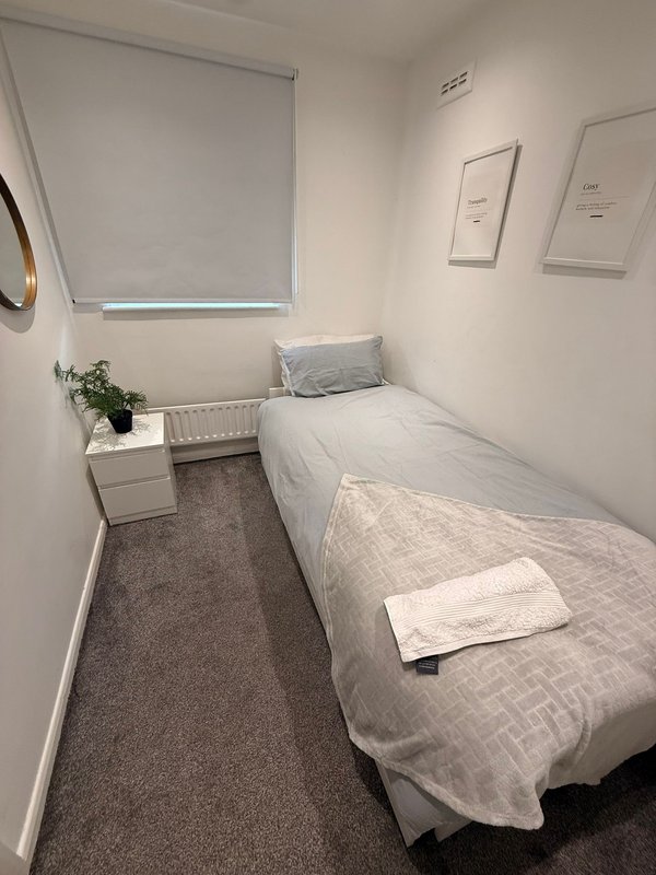 Compact bedroom with single bed, grey carpet, white walls and window blind.