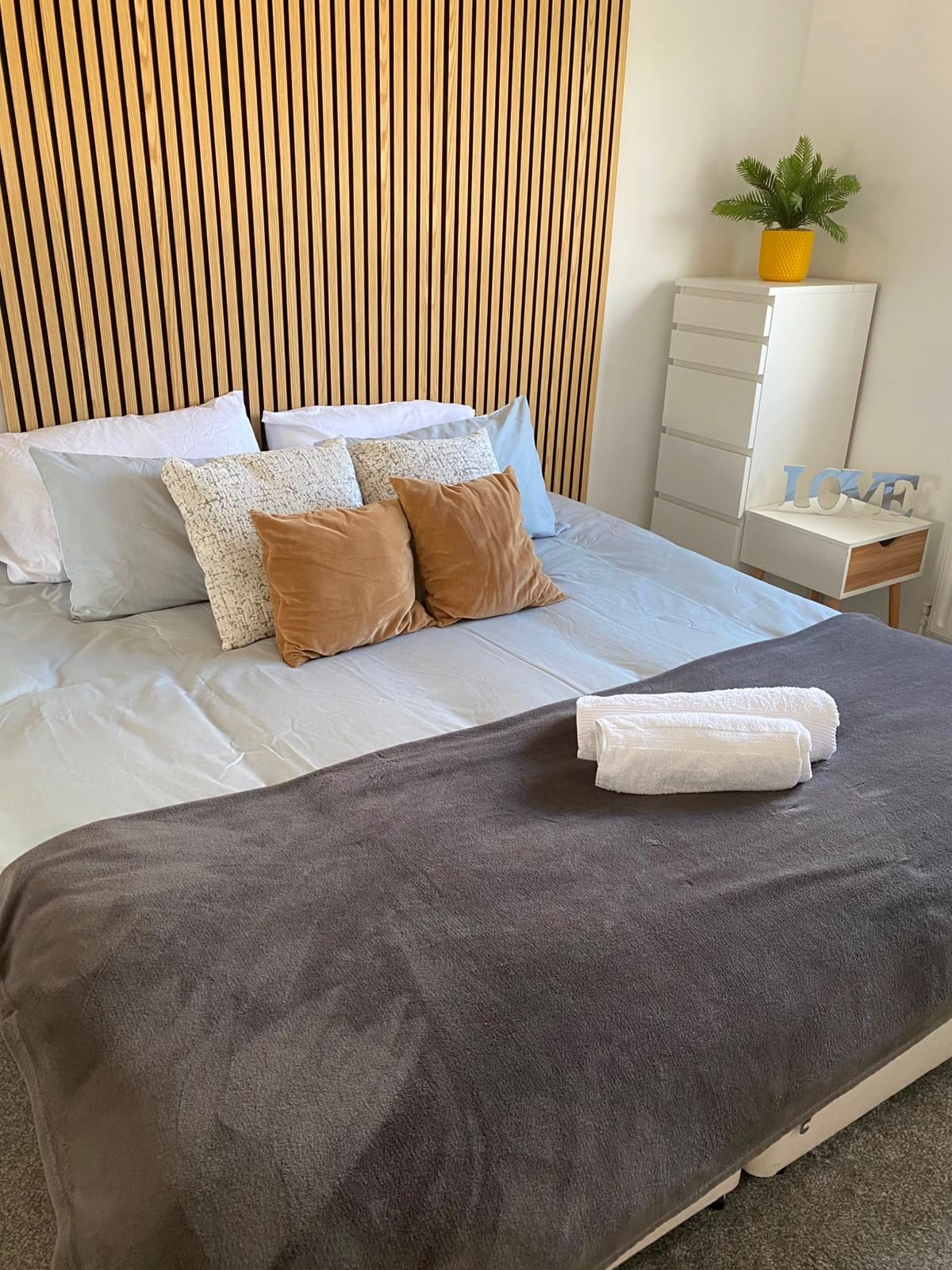 Modern bedroom with double bed, decorative pillows, wooden slat headboard, and potted plant on nightstand.