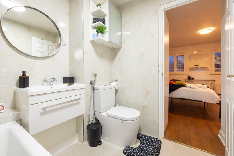 Modern bathroom with white vanity, toilet, round mirror, and view into bedroom beyond