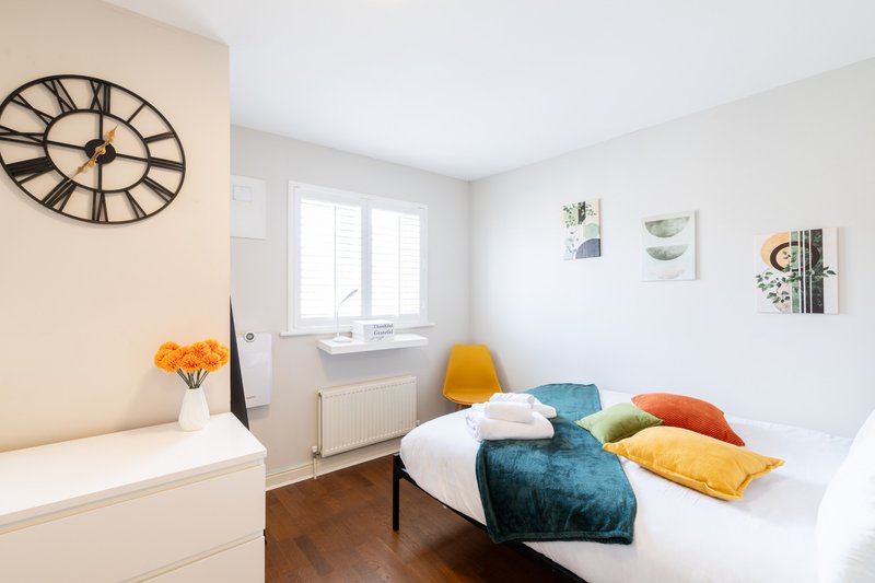 Bright bedroom with double bed, colorful cushions, large wall clock, and window with natural light