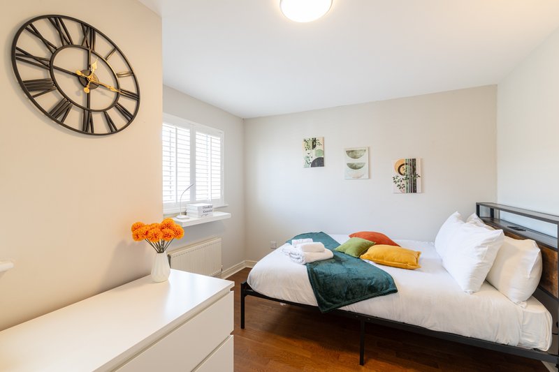 Bright bedroom with double bed, decorative wall clock, window with shutters, and colorful cushions