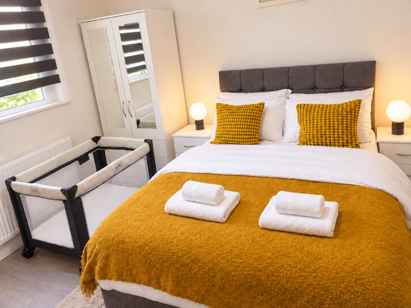 Bedroom with double bed, yellow bedding, gray headboard, folded towels, and travel cot
