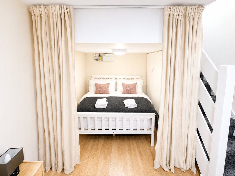 Compact bedroom with white bed frame, black duvet, wooden flooring and cream curtains