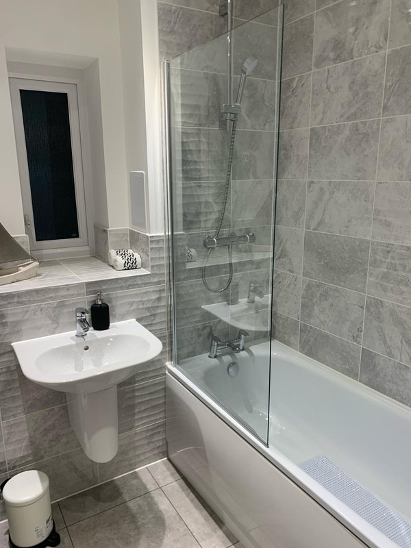 Modern bathroom with grey tile walls, white pedestal sink, bathtub with glass shower screen and toilet