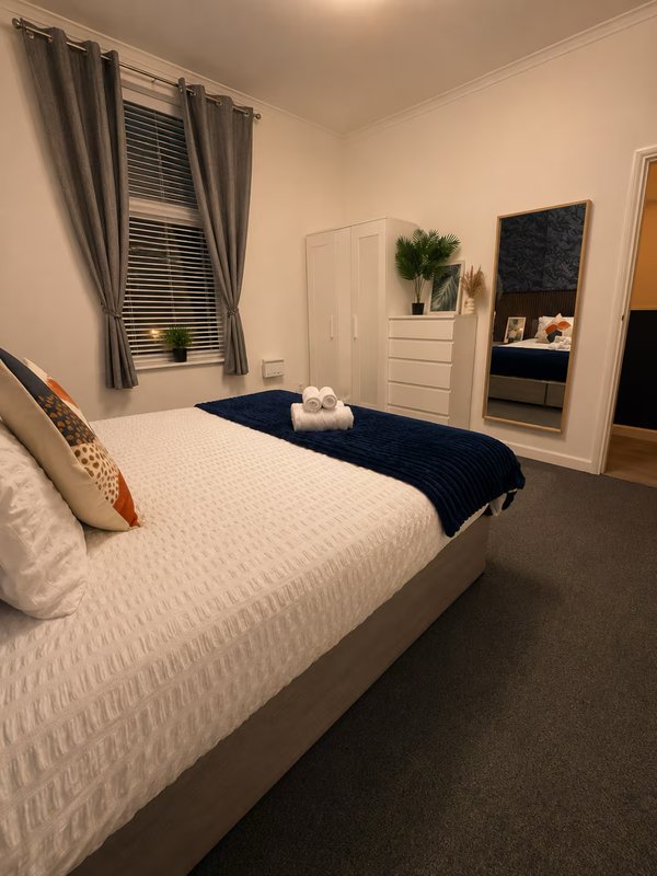 Bedroom with double bed, dark blue throw, white furniture, window with grey curtains