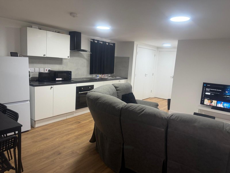 Open-plan kitchen and living room with white cabinets, black sofa, wooden flooring and wall-mounted TV