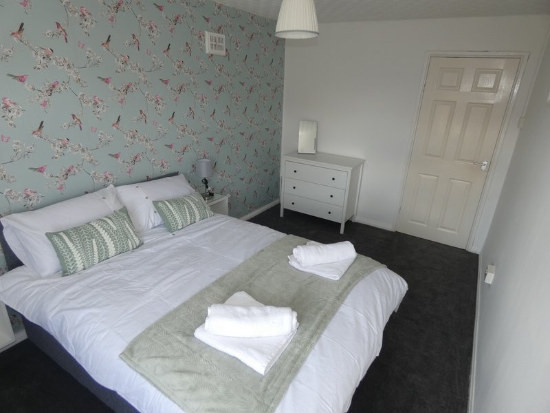 Bright bedroom with double bed, floral wallpaper, white dresser and dark flooring