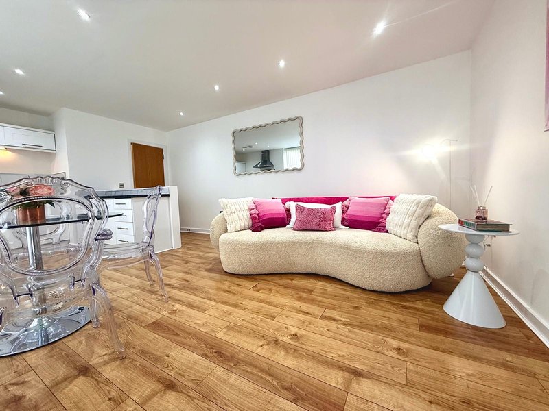 Modern living room with curved cream sofa, pink cushions, wooden floor and kitchenette