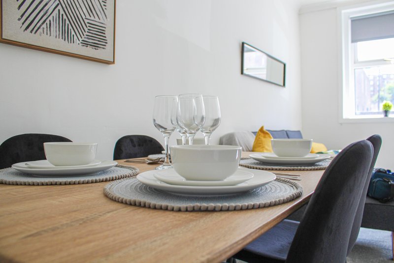 Wooden dining table set with white tableware, glasses, and black chairs near window