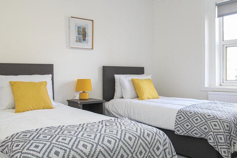 Twin bedroom with two single beds, yellow cushions, grey patterned throws, and window with natural light