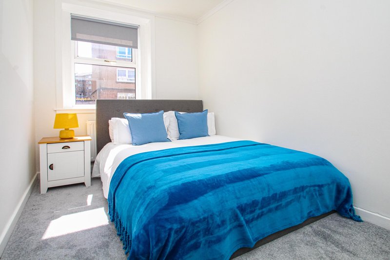 Bright bedroom with double bed, blue bedding, white nightstand with yellow lamp, and window