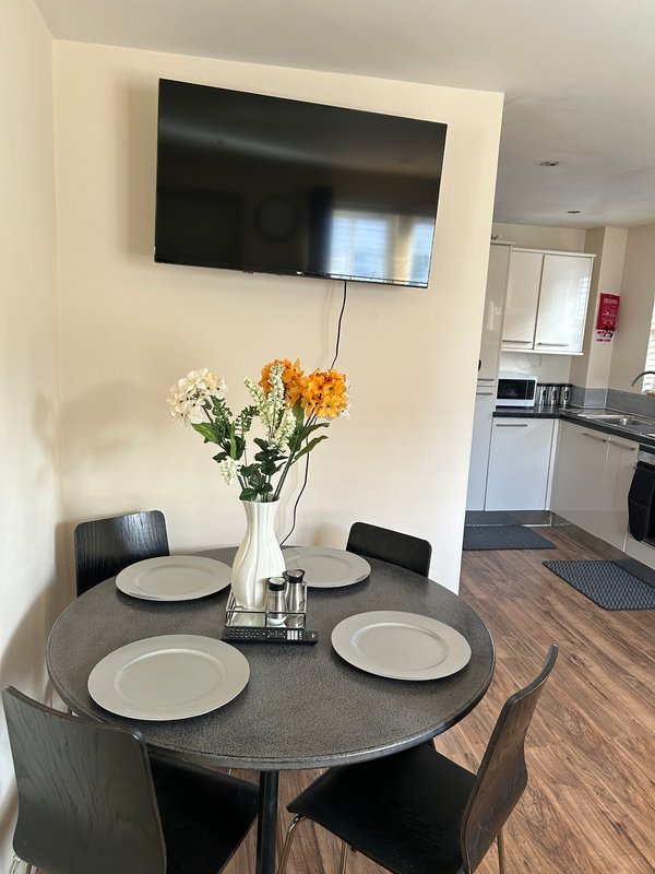 Dining area with round table, place settings, flower vase, wall-mounted TV, and kitchen visible in background