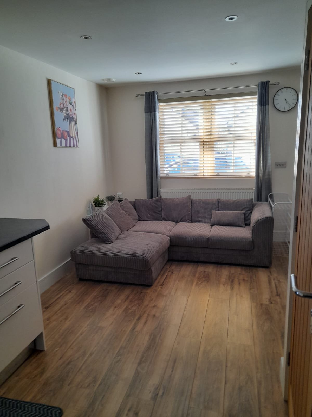 Living room with grey corner sofa, wooden floor, window with blinds and wall clock