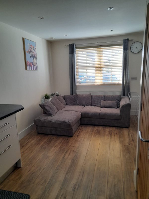 Living room with grey corner sofa, wooden floor, window with blinds and wall clock