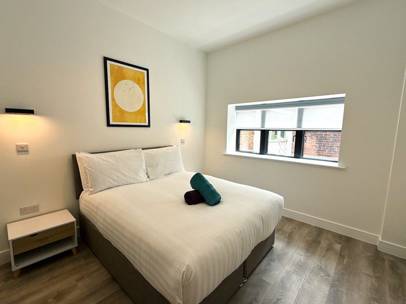 Modern bedroom with double bed, white bedding, dark teal cushion, framed yellow artwork, bedside lamp and window.