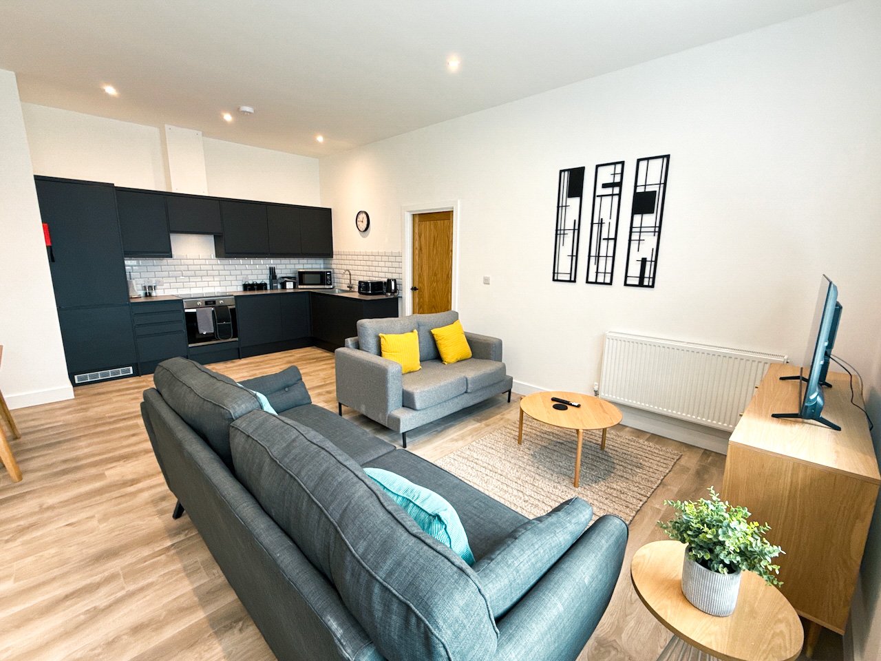 Open-plan living room with dark sofa, kitchen area, yellow cushions, wooden flooring and wall art
