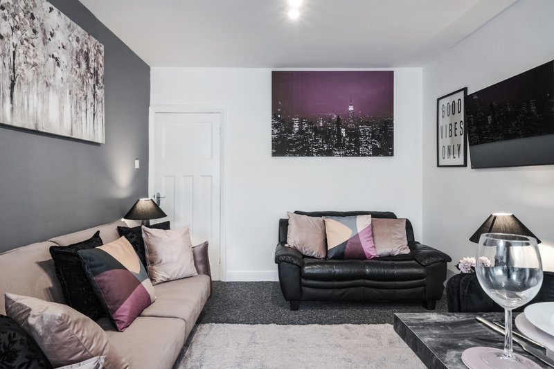 Modern living room with beige sofa, black leather seating, purple and pink cushions, wall art and pendant lighting