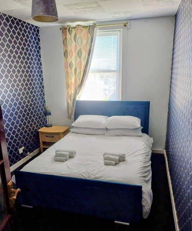 Compact bedroom with double bed, blue headboard, wooden nightstand and patterned curtains