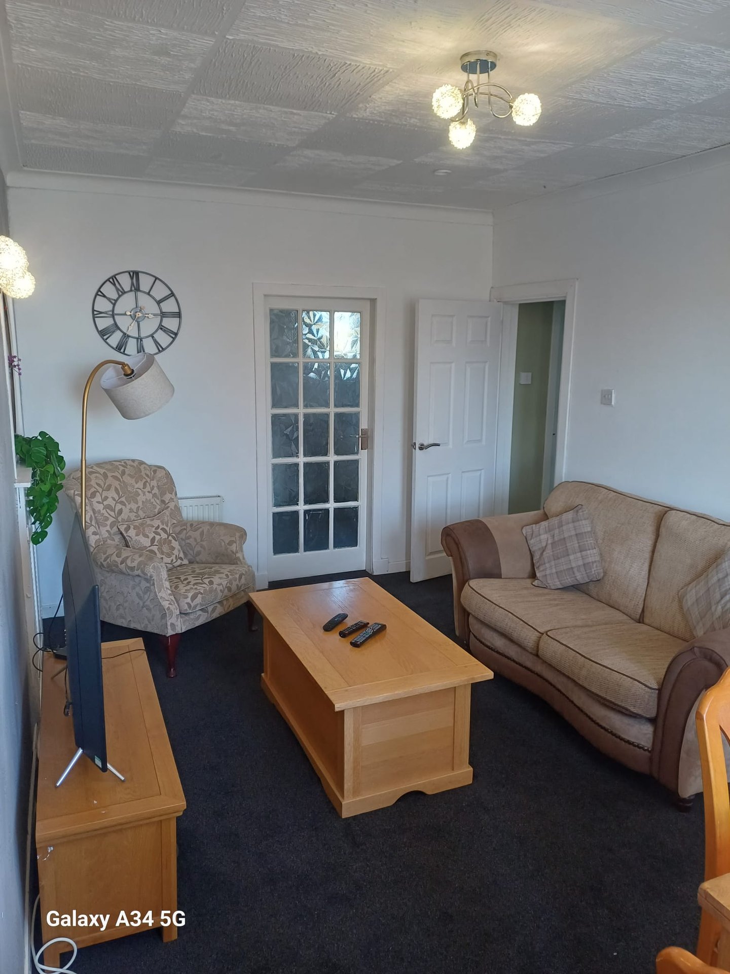 Living room with beige sofa, patterned armchair, wooden coffee table, wall clock and ceiling lights