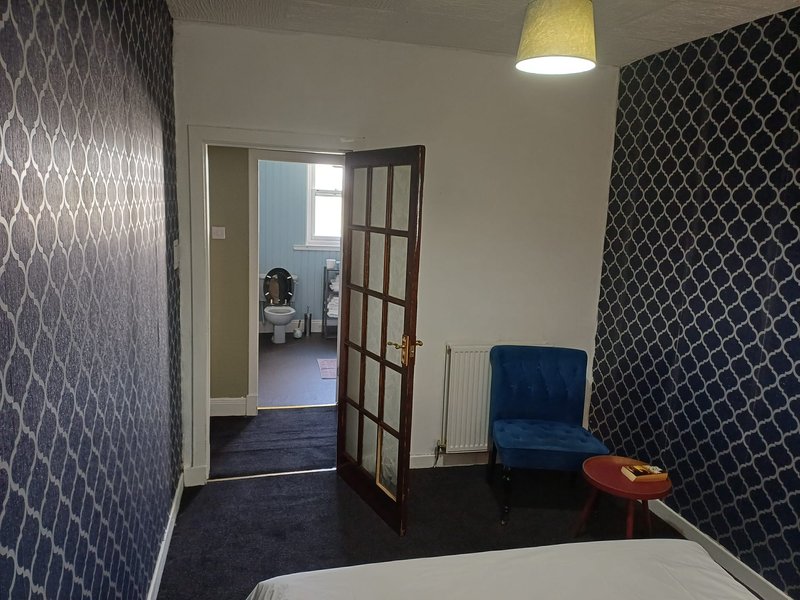 Modern bedroom with patterned wallpaper, blue armchair, open doorway to ensuite bathroom