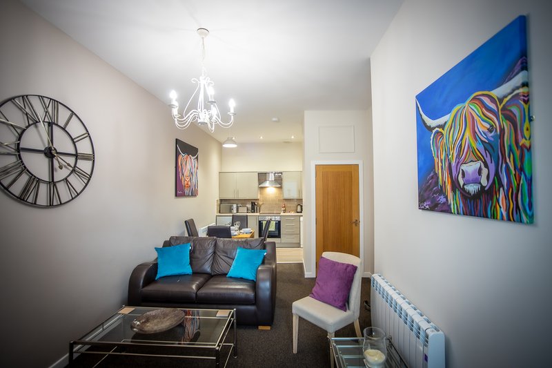 Modern living room with black sofa, blue cushions, colorful wall art, and kitchen visible beyond