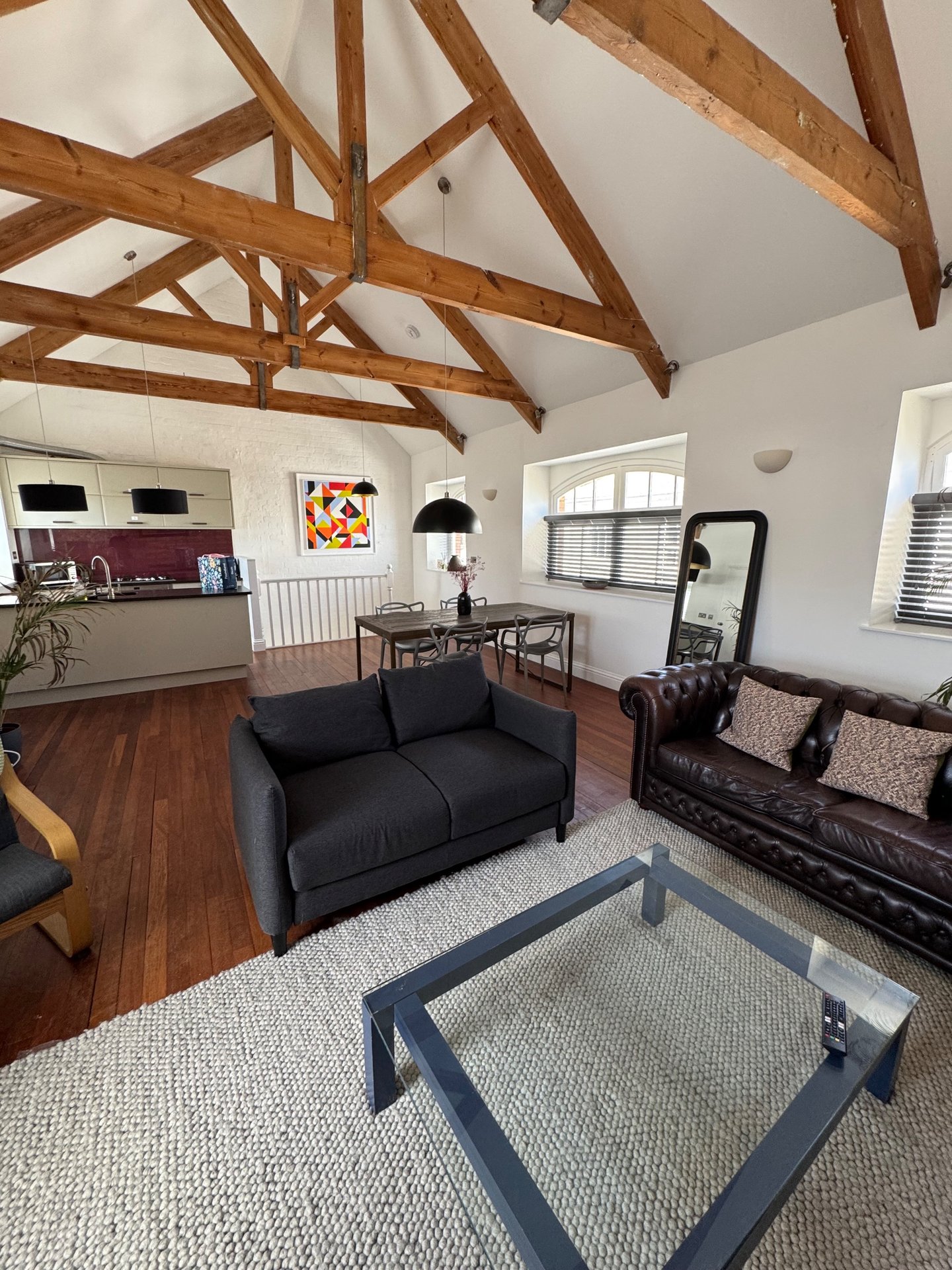 Open-plan living room with exposed wooden beams, black sofa, dining table, and kitchen area
