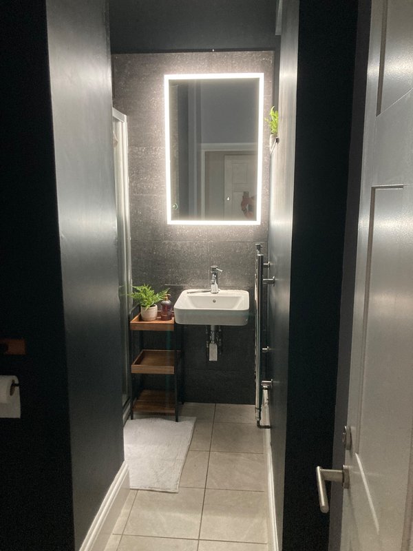 Modern bathroom with illuminated mirror, black vanity, potted plants, and light tile flooring