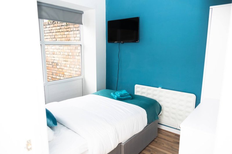 Bedroom with single bed, white bedding, turquoise accent wall, wall-mounted TV, and window